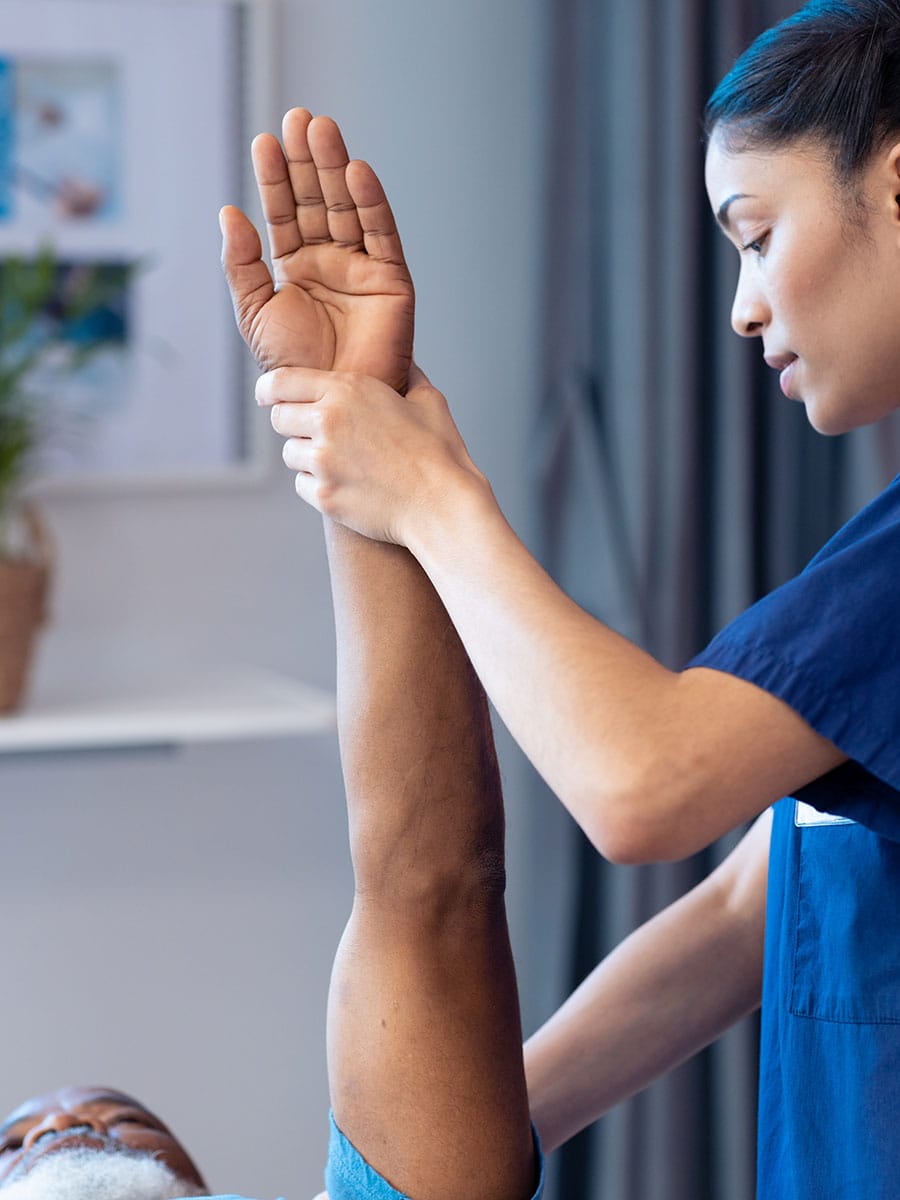 A physical therapist or nurse in blue scrubs performing a guided arm stretch for an elderly male patient.