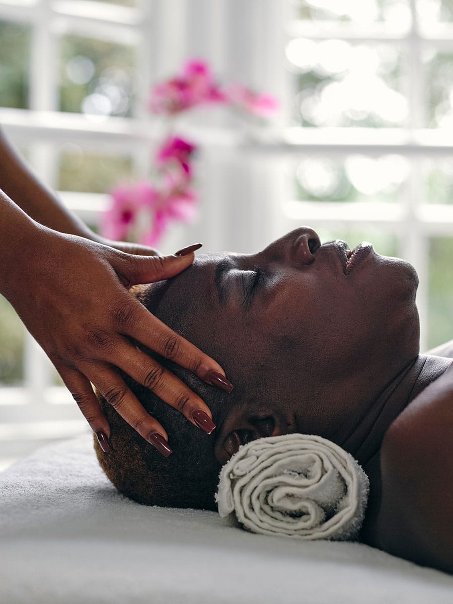 Profile view of a relaxed woman lying down with eyes closed while receiving a gentle head and temple massage from a therapist.