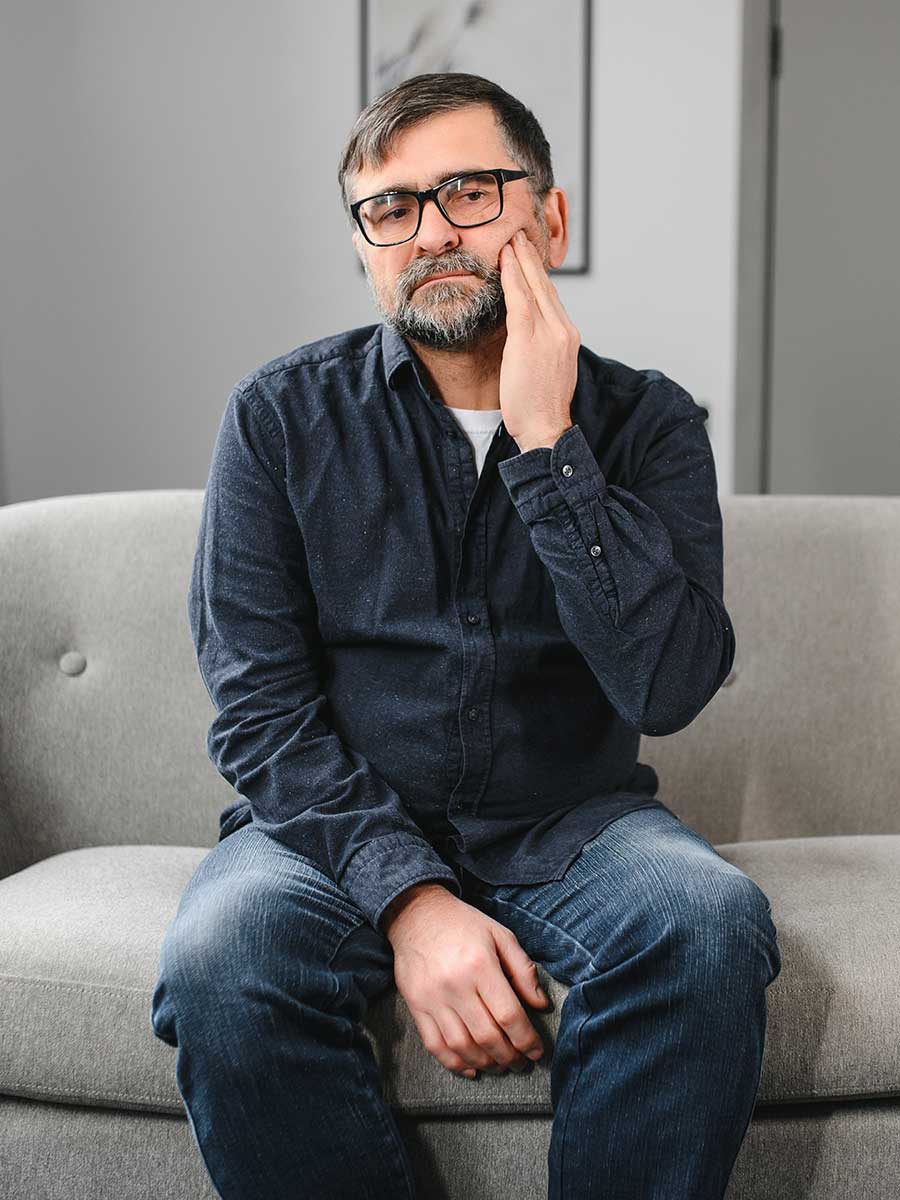 A middle-aged man with glasses and a beard sitting on a grey sofa, holding his hand against his cheek with an uncomfortable expression.