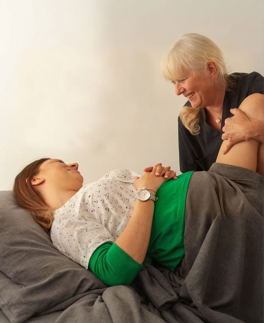 A smiling blonde female osteopath or massage therapist gently treating a patient's leg while the patient lies comfortably on a treatment table.