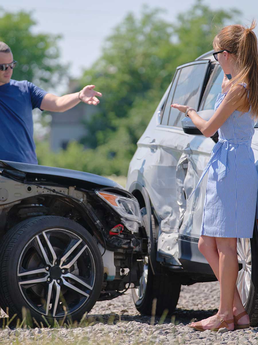 A man and a woman standing next to two collided cars on the side of a road, gesturing in distress after a car accident.