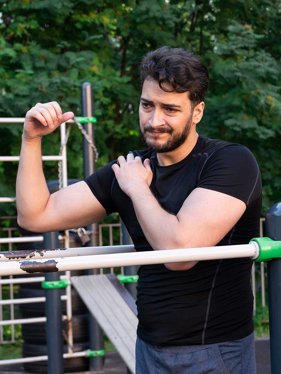 A young man with a beard leaning against parallel bars at an outdoor gym, gripping his shoulder with a pained expression.