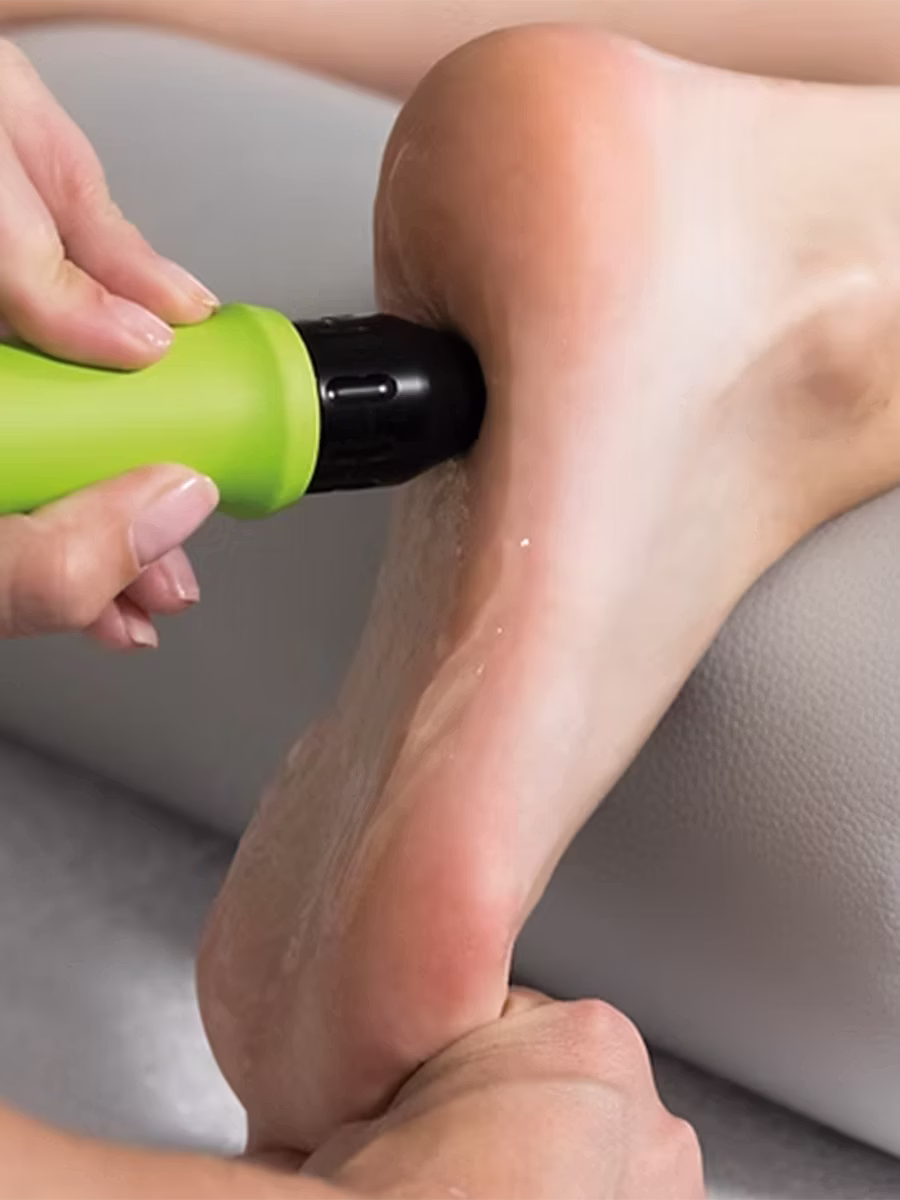 A close-up view of a medical professional's hands applying shockwave therapy to a patient's heel and foot. The clinician holds the end of a lime green and black therapy device against the patient's skin, which is covered in a clear ultrasound gel. The patient's foot rests on a white leather treatment table.
