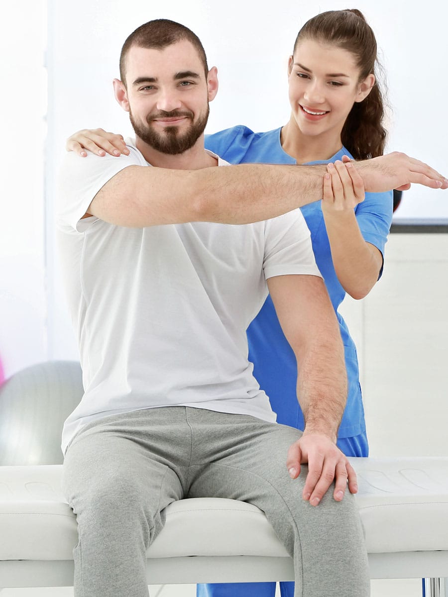 A smiling man with a beard sits on a white treatment table while a female practitioner in blue scrubs assists him with a cross-body arm stretch. The practitioner stands behind him, guiding his arm across his chest by holding his wrist and shoulder. The background is a bright, clinical setting with a fitness ball visible.