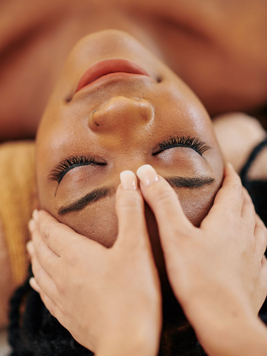 A woman with her eyes closed and a peaceful expression receiving a forehead massage from a therapist.