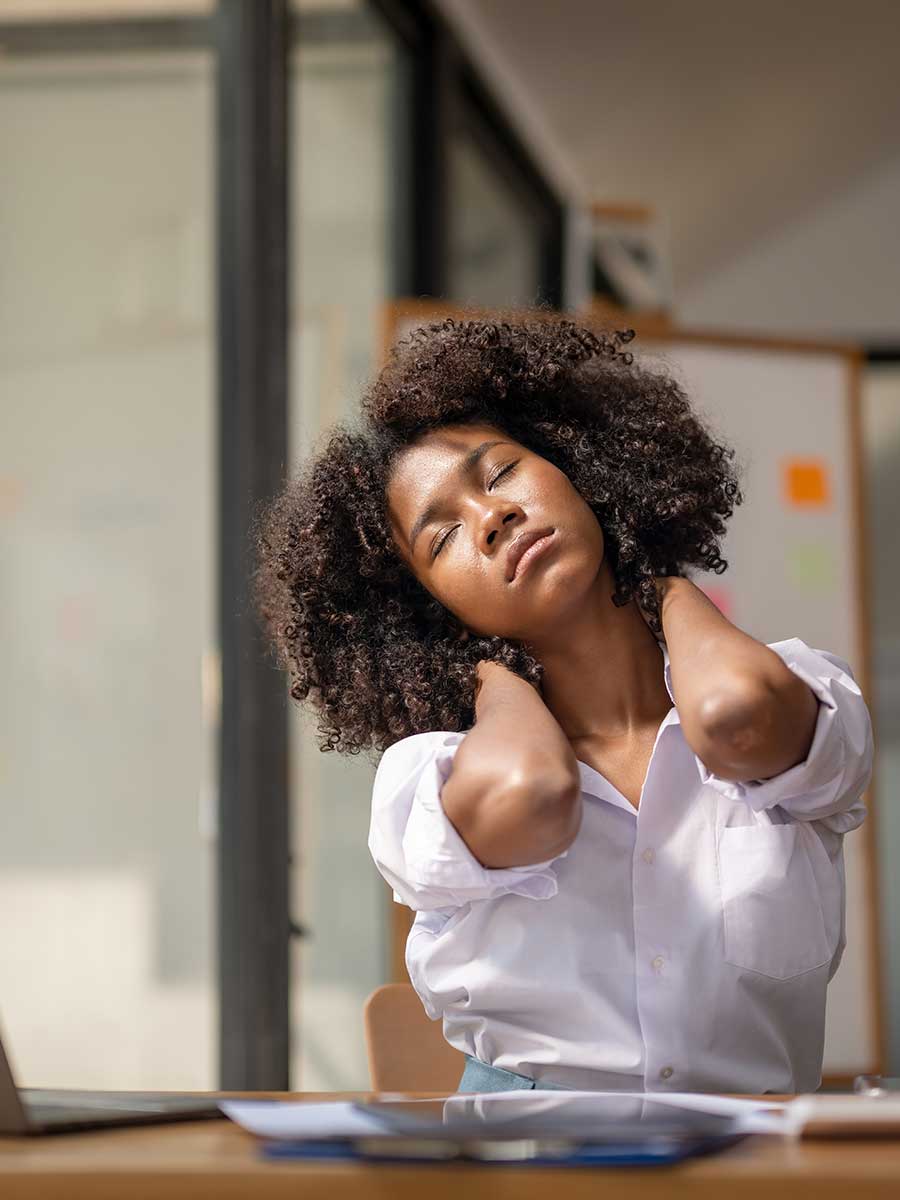 A woman with curly hair sitting at an office desk with her eyes closed, holding the back of her neck and stretching her neck muscles to relieve tension.